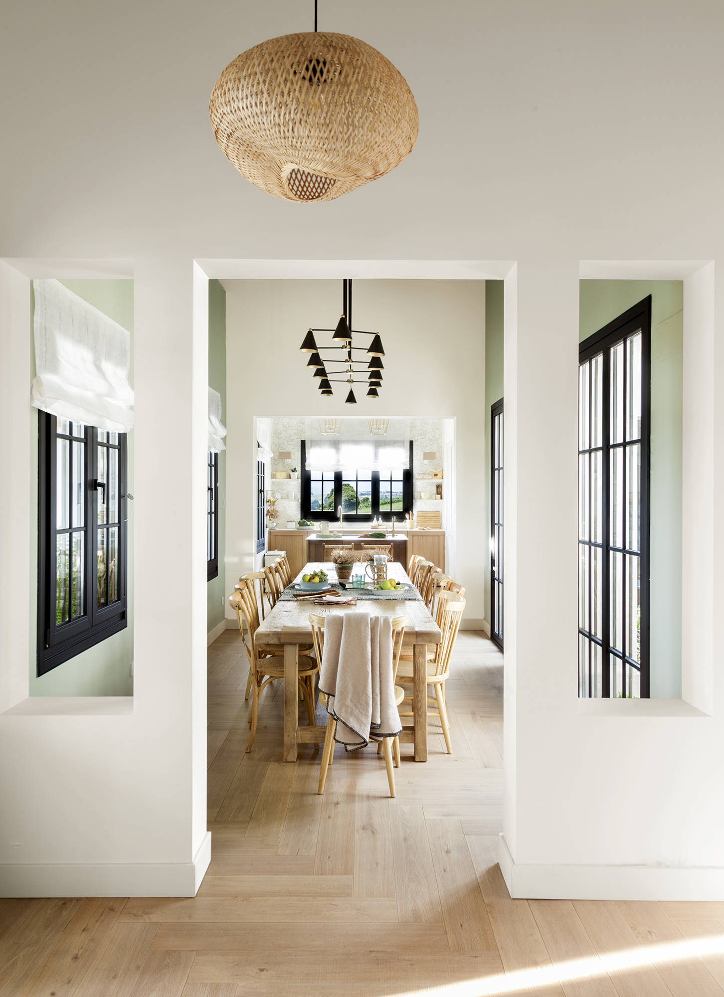 Comedor con mesa de madera, ventanales negros, lámpara de fibras y negra y cocina al fondo. 